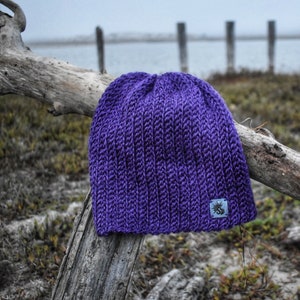 May include: Purple knitted beanie with a small logo on the side. The beanie is sitting on a piece of driftwood.