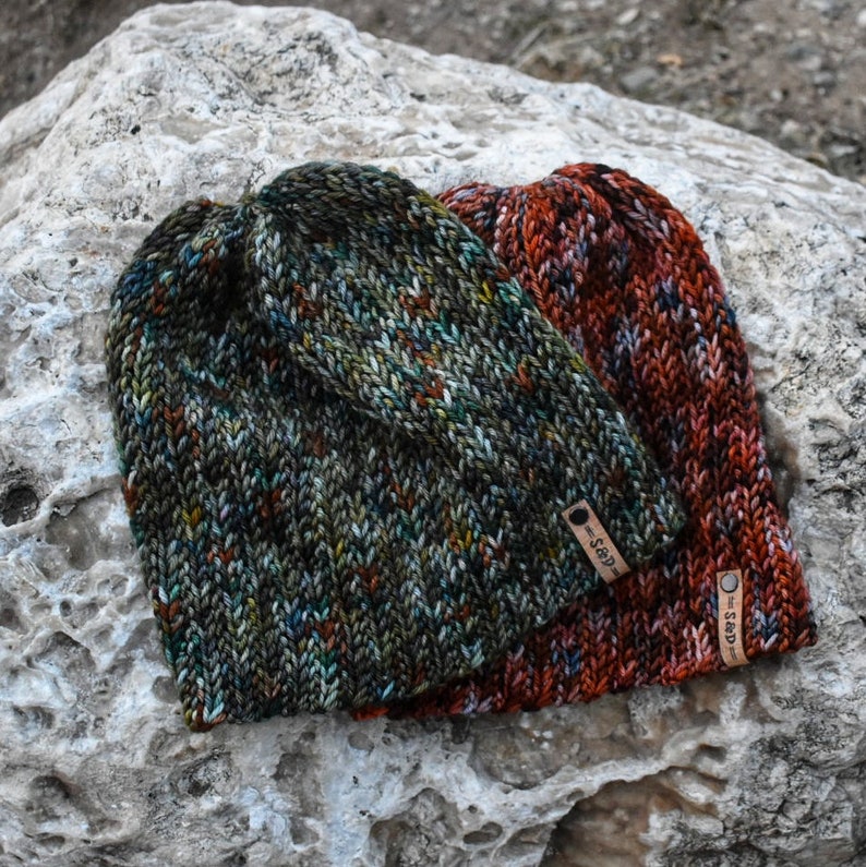 May include: Two knitted hats with a colorful, speckled pattern. The hats are made with a soft yarn and have a unique, hand-crafted look. The hats have leather tags with the text "S&D" on them.