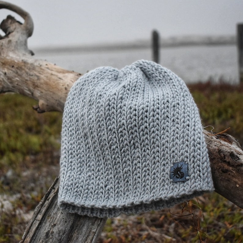 May include: A gray knitted beanie with a small, dark gray patch on the side. The beanie is sitting on a piece of driftwood.