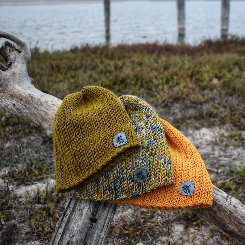 May include: Three knitted hats in different colors and patterns. The hats are stacked on top of each other and have small leather tags with a sun design.