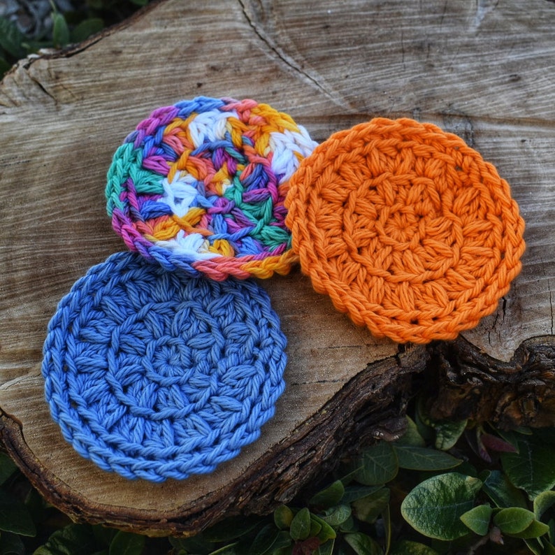 May include: Three crocheted cotton rounds in different colours: blue, orange, and multi-coloured. The rounds are on a wooden surface.