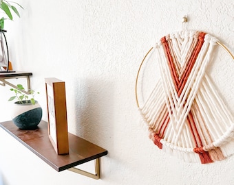 May include: A macrame wall hanging with a circular wooden frame and a geometric pattern of white, peach, and terracotta yarn. The hanging has a fringe of yarn at the bottom.