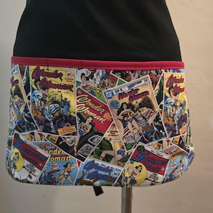 May include: Black apron with a Wonder Woman comic book print on the lower half. The apron has a red trim and a black waistband. The comic book print features various Wonder Woman comic book covers.