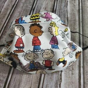 May include: A white fabric face mask with a colorful Peanuts cartoon print featuring characters like Charlie Brown, Snoopy, Lucy, and Linus.