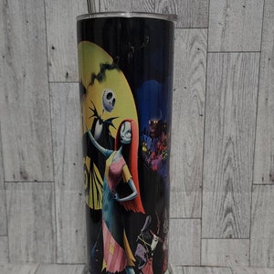 Puede incluir: Un vaso de acero inoxidable blanco y negro con una imagen colorida de Jack Skellington y Sally de la pel&iacute;cula Pesadilla antes de Navidad. El vaso tiene una tapa plateada y una pajita.