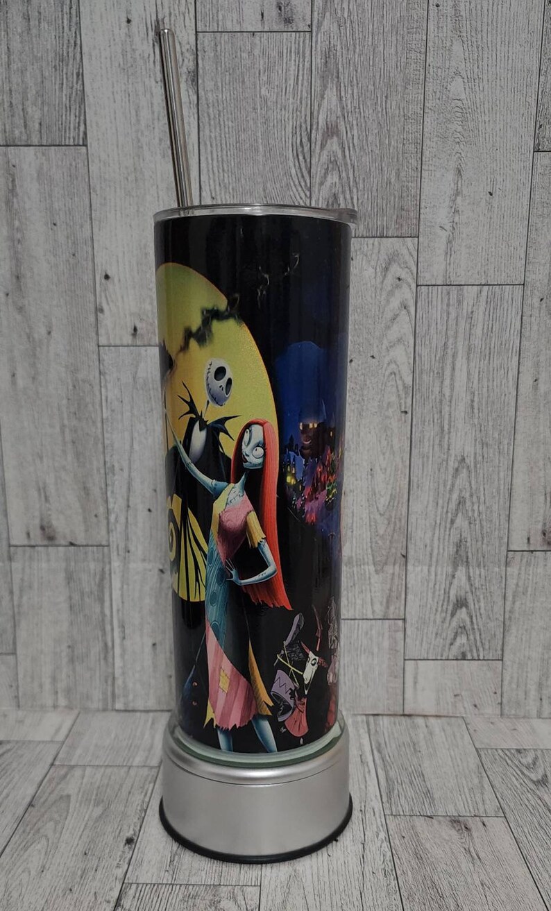 Puede incluir: Un vaso de acero inoxidable blanco y negro con una imagen colorida de Jack Skellington y Sally de la pel&iacute;cula Pesadilla antes de Navidad. El vaso tiene una tapa plateada y una pajita.