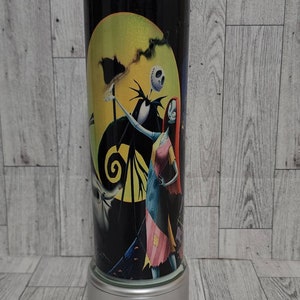 Puede incluir: Un vaso negro y blanco con una imagen colorida de Jack Skellington y Sally de Pesadilla antes de Navidad. El vaso tiene una base plateada y una pajita transparente.