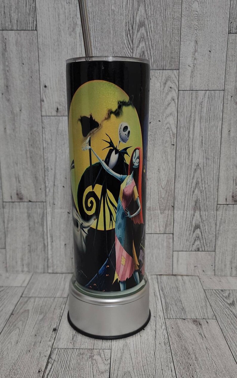 Puede incluir: Un vaso negro y blanco con una imagen colorida de Jack Skellington y Sally de Pesadilla antes de Navidad. El vaso tiene una base plateada y una pajita transparente.