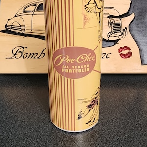 May include: A yellow tumbler with brown vertical stripes and vintage illustrations. The tumbler features the text "Pee-Chee ALL SEASON PORTFOLIO" in a maroon oval. A wooden sign with a car silhouette is in the background.