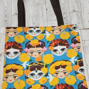 May include: A blue tote bag with a colorful print of Day of the Dead inspired faces with marigold flowers. The bag has brown straps.