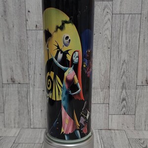 Puede incluir: Un vaso negro y plateado con un gr&aacute;fico de Jack Skellington y Sally de Pesadilla antes de Navidad. El vaso tiene una tapa plateada y una pajita.