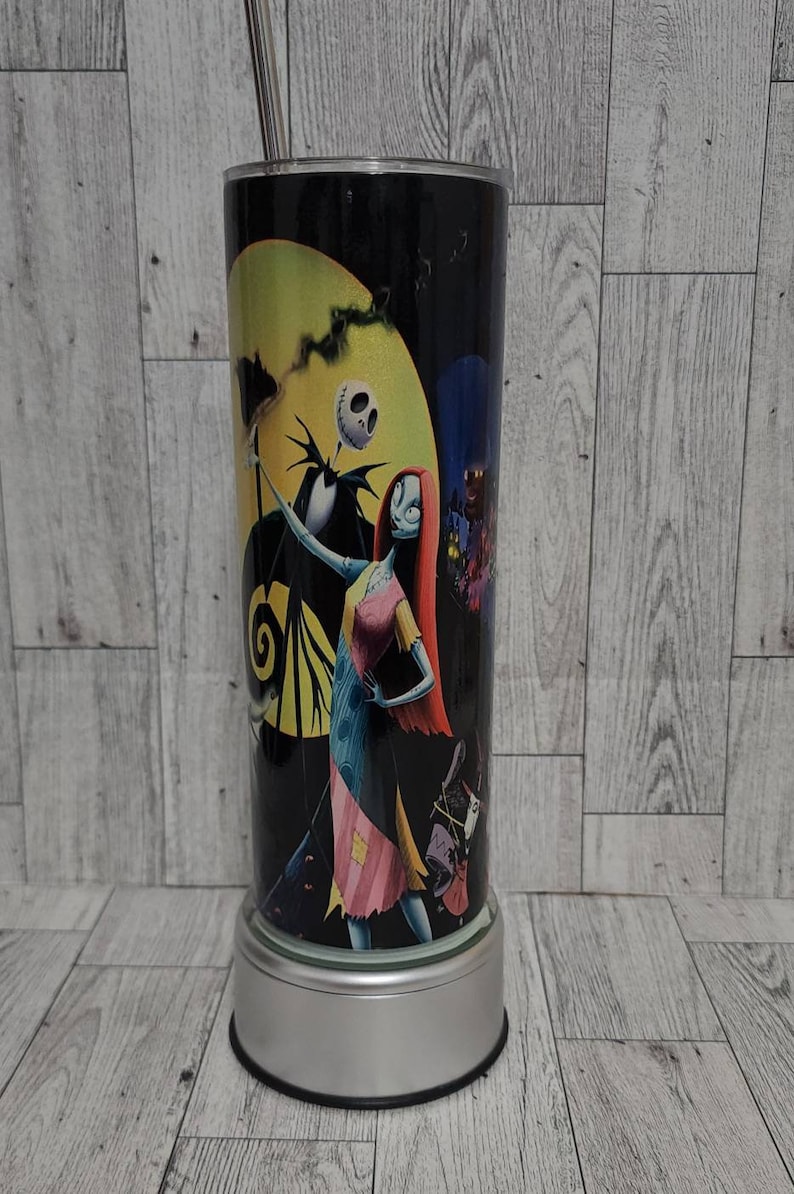 Puede incluir: Un vaso negro y plateado con un gr&aacute;fico de Jack Skellington y Sally de Pesadilla antes de Navidad. El vaso tiene una tapa plateada y una pajita.