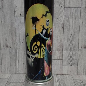 Puede incluir: Un vaso de acero inoxidable blanco y negro con una imagen colorida de Jack Skellington y Sally de la pel&iacute;cula Pesadilla antes de Navidad. El vaso tiene una tapa plateada y una base plateada.