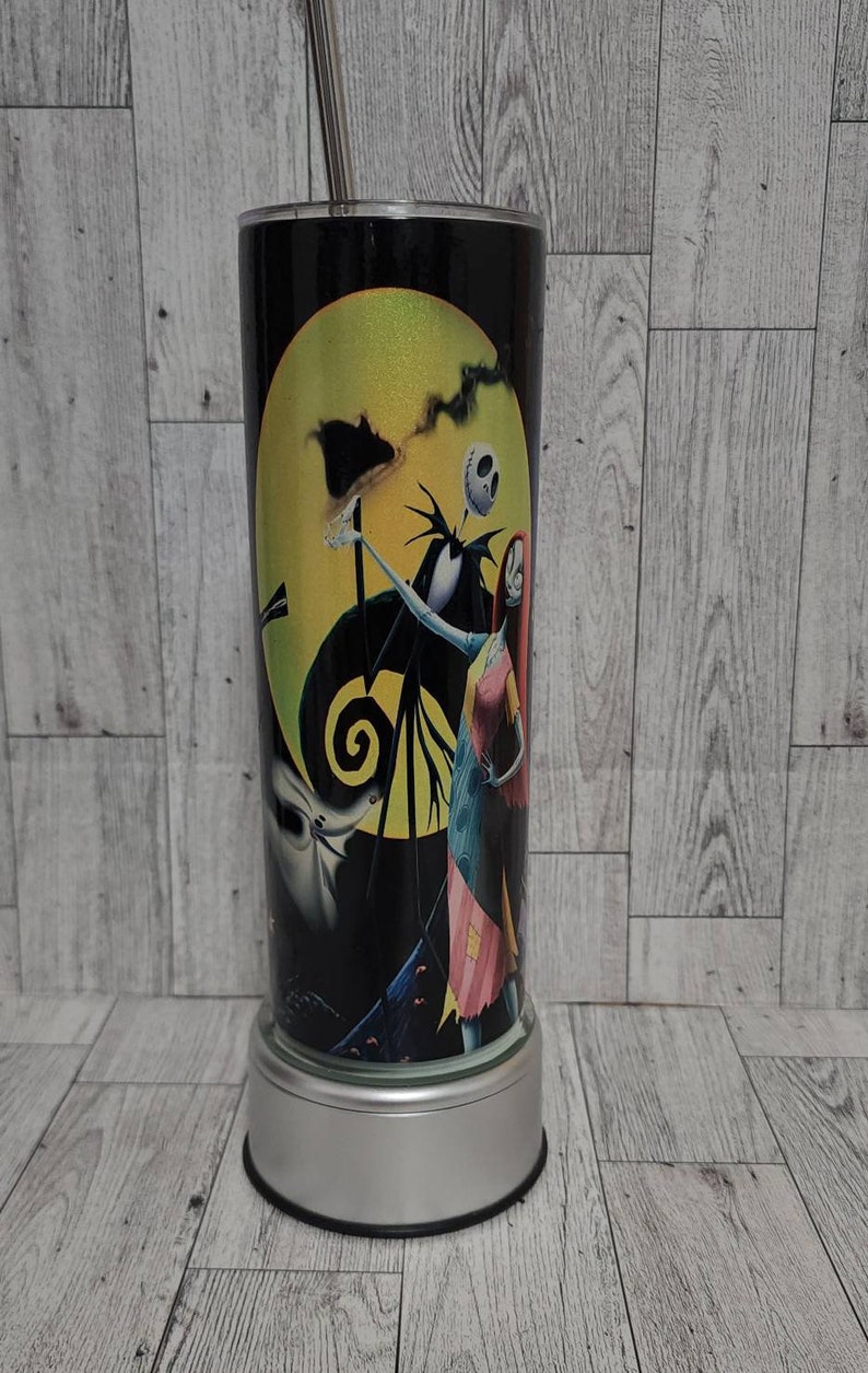 Puede incluir: Un vaso de acero inoxidable blanco y negro con una imagen colorida de Jack Skellington y Sally de la pel&iacute;cula Pesadilla antes de Navidad. El vaso tiene una tapa plateada y una base plateada.