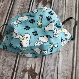 May include: A blue fabric face mask with a repeating pattern of Snoopy and Woodstock characters, paw prints, and flowers. The mask has black elastic ear loops.