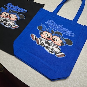 May include: Two tote bags, one black and one blue, featuring a graphic of Mickey and Minnie Mouse in baseball uniforms. The word "Dodgers" is printed above the characters in a script font. Both bags have black handles.