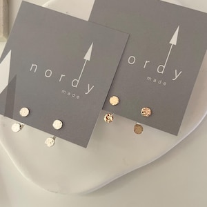 May include: Two pairs of small, hammered, round earrings. One pair is silver and the other is gold. The earrings are displayed on a white surface with two gray cards. The cards have the text "nordy made" on them.