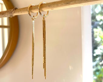 May include: A pair of gold dangle earrings with a chain design. The earrings are hanging from a wooden rod.