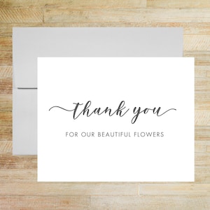 May include: A white folded greeting card with a matching white envelope. The front of the card features the dark gray text thank you in a cursive script font, followed by the text FOR OUR BEAUTIFUL FLOWERS in a simple sans-serif font.