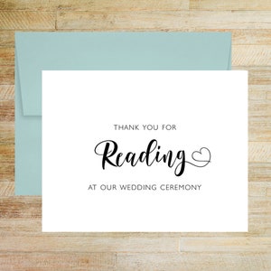 May include: A white card with the words "Thank You For Reading at Our Wedding Ceremony" in black script. A light blue envelope is behind the card. The card has a heart symbol after the word "Reading".