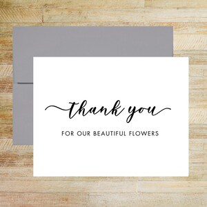 May include: A white rectangular greeting card with a grey envelope. The card features the black text "thank you" in a cursive script font, followed by the text "FOR OUR BEAUTIFUL FLOWERS" in a simple, clean, black sans-serif typeface.