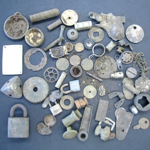 May include: An assortment of small, weathered metal objects, including gears, padlocks, and various hardware pieces. The collection features a range of shapes and sizes, with a muted, aged appearance. The items are scattered on a dark surface.