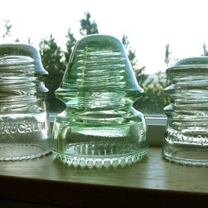 May include: Three antique glass insulators in shades of green and clear sit on a wooden surface. The center insulator is a vibrant green, while the others are clear. The word "MCLAUGHLIN" is visible on one.