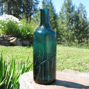 May include: A vintage teal-colored glass bottle with the embossed word "Palmer" on the side. The bottle is cylindrical with a long neck and sits on a stone surface. The background features green grass, trees, and a bright blue sky.