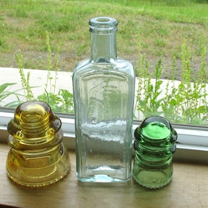 May include: A clear glass bottle with a rectangular body and a narrow neck, flanked by two glass insulators. On the left is an amber-colored glass insulator, and on the right is a smaller, vibrant green glass insulator with a rounded top.