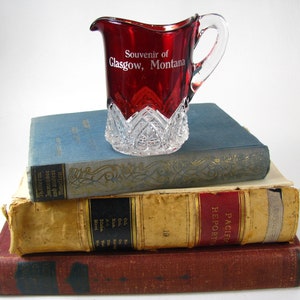 May include: A red glass pitcher with a clear glass base and a white inscription that reads "Souvenir of Glasgow, Montana". The pitcher is sitting on top of a stack of three books.