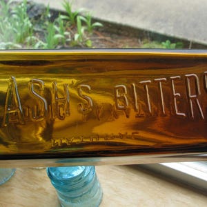 May include: A rectangular amber glass bottle with the embossed text "LASH'S BITTERS" in white lettering. The bottle is positioned horizontally, with the text clearly visible. The background includes a window and other bottles.