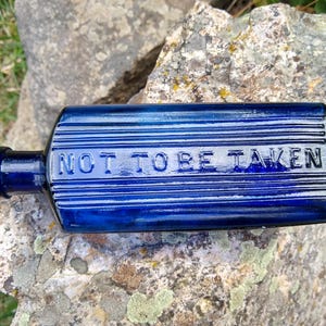 May include: A cobalt blue antique glass bottle with the embossed text "NOT TO BE TAKEN". The rectangular bottle has horizontal ridges and a rounded neck. The bottle is displayed on a textured rock surface.