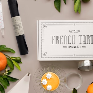 May include: A baking kit for French tarts, including a box with the text "FRENCH TARTS", a perforated baking mat, and a tart ring. The image also features a thermometer, piping bag, and a finished tart with white frosting. Oranges and green leaves are scattered around the items.