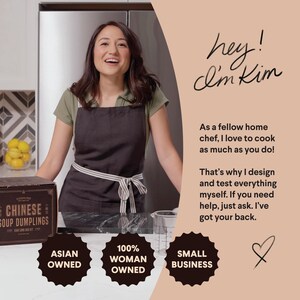 May include: A woman wearing a black apron stands in a kitchen holding a box of Chinese soup dumplings. The box has the text "Cooking Joy" and "Chinese Soup Dumplings" on it. The woman is smiling and looking at the camera. The text "hey! I'm Kim" is written in cursive on a peach background. The text "As a fellow home chef, I love to cook as much as you do!" is written below the cursive text. The text "That's why I design and test everything myself. If you need help, just ask. I've got your back." is written below the previous text. Three black and white circles with the text "Asian Owned", "100% Woman Owned", and "Small Business" are on the counter in front of the woman.