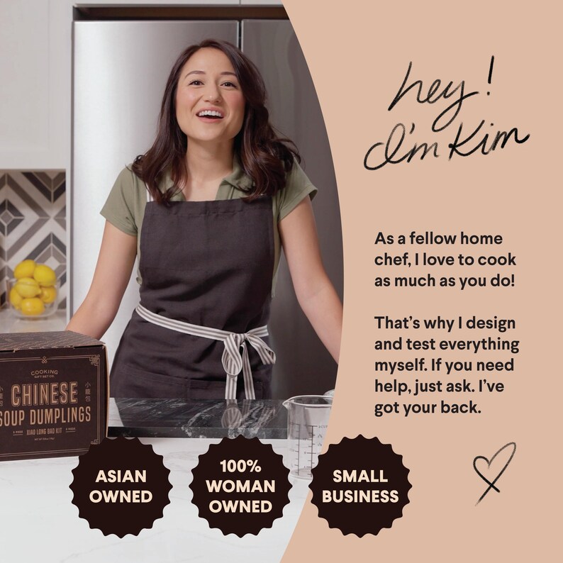 May include: A woman wearing a black apron stands in a kitchen holding a box of Chinese soup dumplings. The box has the text "Cooking Joy" and "Chinese Soup Dumplings" on it. The woman is smiling and looking at the camera. The text "hey! I'm Kim" is written in cursive on a peach background. The text "As a fellow home chef, I love to cook as much as you do!" is written below the cursive text. The text "That's why I design and test everything myself. If you need help, just ask. I've got your back." is written below the previous text. Three black and white circles with the text "Asian Owned", "100% Woman Owned", and "Small Business" are on the counter in front of the woman.