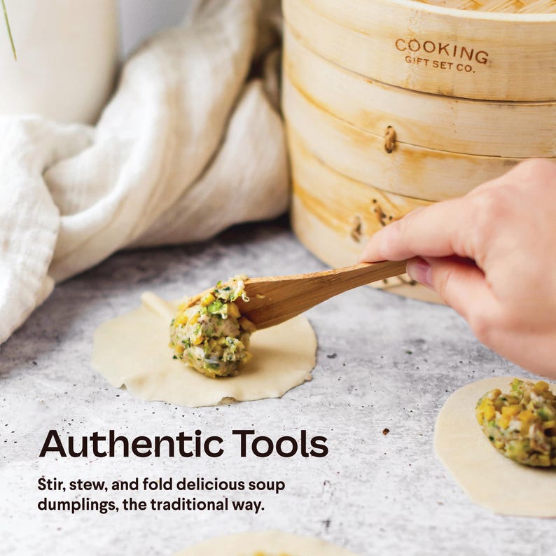 May include: A wooden spoon is used to fill a round dumpling wrapper with a mixture of vegetables and meat. The dumpling is on a gray surface. The text "Authentic Tools" is below the dumpling. The text "Stir, stew, and fold delicious soup dumplings, the traditional way." is below the text "Authentic Tools". The text "COOKING GIFT SET CO." is on a wooden steamer in the background.