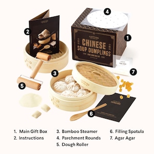 May include: A Chinese soup dumpling kit with a bamboo steamer, parchment rounds, a dough roller, a filling spatula, and agar agar. The kit comes in a black gift box with gold lettering that reads "Cooking  by the  Sea  Chinese Soup Dumplings  Xiao Long Bao Kit".