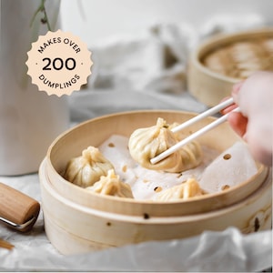 May include: A wooden steamer basket filled with white dumplings. A hand uses chopsticks to pick up a dumpling. The text "Makes Over 200 Dumplings" is on a brown circle.