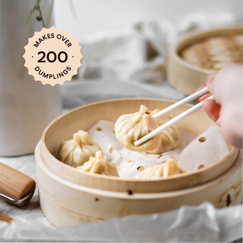 May include: A wooden steamer basket filled with white dumplings. A hand uses chopsticks to pick up a dumpling. The text "Makes Over 200 Dumplings" is on a brown circle.