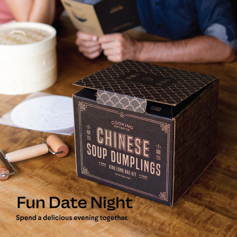 May include: A black box with gold lettering that says "Cooking Gift Set Co. Chinese Soup Dumplings Xiao Long Bao Kit". The box is open and the contents are visible, including a rolling pin and round white paper.