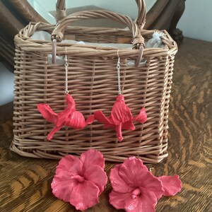 May include: A pair of pink hibiscus flower earrings and two additional pink hibiscus flowers. The earrings are hanging from a wicker basket.