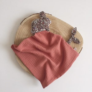 May include: A coral colored muslin baby blanket with a floral print bunny attached. The bunny has a bow on its tail.