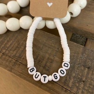 May include: White beaded bracelet with the words "BUT GOD" spelled out in black letters.
