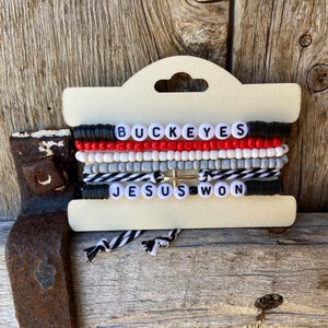 May include: A set of five beaded bracelets with the words "BUCKEYES" and "JESUS WON" spelled out in black letters. The bracelets are on a wooden display card with a hole for hanging.