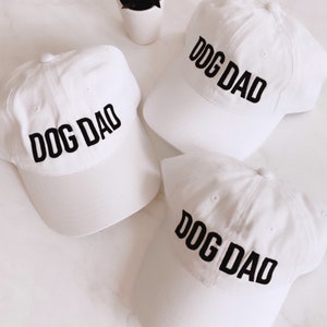 May include: Three white baseball caps with black embroidered text that reads "DOG DAD".
