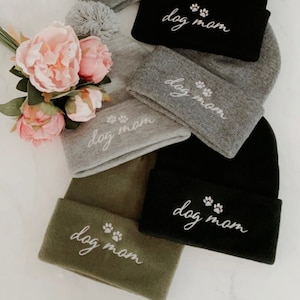 May include: A collection of beanies in various colors, including gray, black, and olive green. Each beanie features the embroidered text "dog mom" with paw prints. Some beanies have a matching pom-pom on top. Pink flowers are in the background.