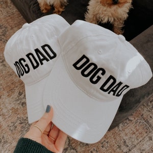 May include: Two white baseball caps with black text that reads "Dog Dad" on each. The caps are on a light brown surface.