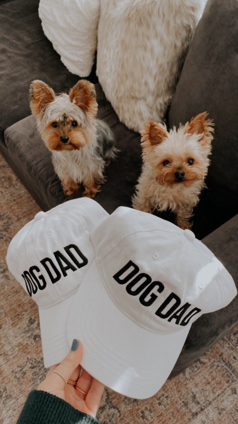 May include: Two white baseball caps with black text that reads "Dog Dad" on each. The caps are on a light brown surface.