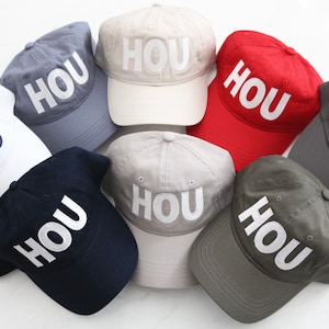 May include: A collection of baseball caps in various colors, including white, navy, gray, red, and beige. Each cap features the word "HOU" in bold, white lettering across the front. The caps are arranged in a circular pattern.