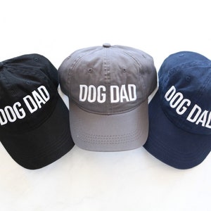 May include: Three baseball caps in black, gray, and navy blue with the text "DOG DAD" embroidered in white on the front of each cap.